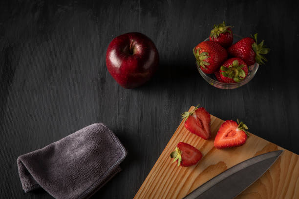 Strawberries in a glass container on a wooden table, with a cutting board, with cut strawberries and a knife next to it, accompanied by a red apple.