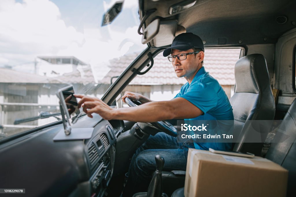 asian chinese mature male delivery person using digital tablet mobile app checking delivery information in driver's seat in delivery van Truck Driver Stock Photo