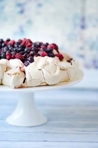 Berries and cream atop a pavlova on a cake stand with a blue background.