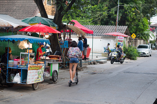 Young thai woman is driving on electro scooter in residential area of Ratchada 36 in Bangkok. Woman is passing some street food vendors. SHe is wearing shorts and shirt with Disney characters
