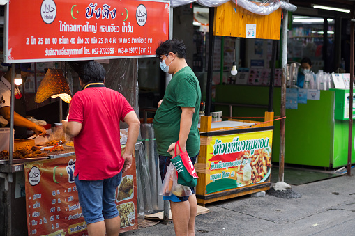 Thai men with masks are buying street food in Bangkok. Scene is close to university CRU in Bangkok. Around gate to university are several food stalls. People are casually dressed