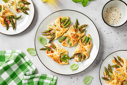 Baked green asparagus with ham and cheese in puff pastry sprinkled with sesame seeds and green basil leaves. Light gray stone background. top view