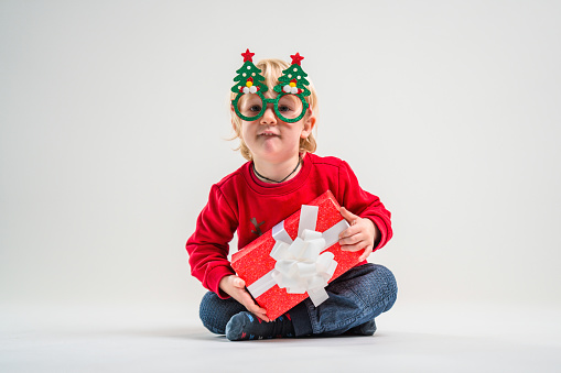 Cute Caucasian blond toddler boy is holding Christmas present and looking at camera. He is wearing Christmas sweater, photo booth eyeglasses and is happy. White background.