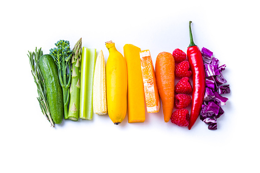 Top view of various fresh fruits and vegetables sticks arranged side by side by colors on a rainbow gradient on white background. The composition includes rosemary, a pickle, broccoli, a asparagus, a celery stick, a tiny sweetcorn, a banana, a pumpkin stick, a sliced grapefruit, a carrot, various raspberries, a red hot chili pepper, and a chopped red cabbage.