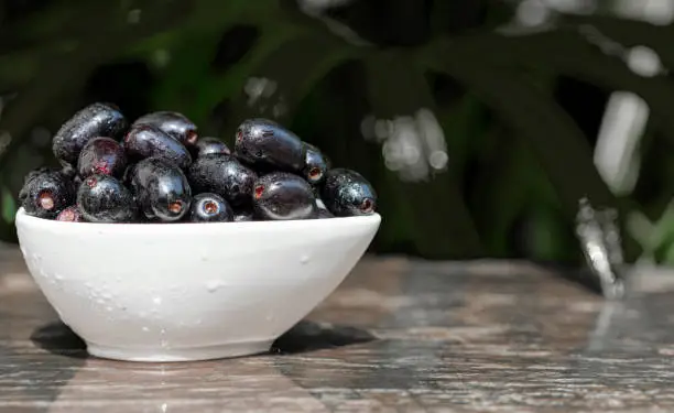 Closeup shot. A cup of fresh Black plums or Java plums in dark purple color with water drops on a wooden table as background. Wild fruits with healthy nutrition