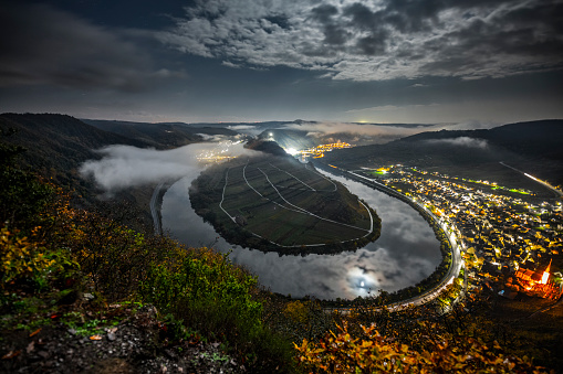 Moonlit Moselle Bend (Moselschleife) at the village Bremm in Rhineland-Palatinate, Germany at night. Moody fog is flowing though the valley.