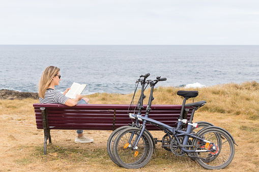 Woman sitting on a bench in front of the sea reading a book and two bikes leaning against the bench.