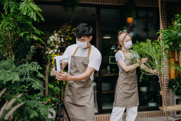 Young Asian couple, running a small business flower shop. Wearing protective face mask to prevent the spread of Coronavirus. Setting up non-contact infrared thermometer scanner preparing to re-start the business. Re-opening after Covid-19 concepts joven pareja asiática, dirigiendo una pequeña floristería de negocios. usar máscara facial protectora para prevenir la propagación del coronavirus. configuración de escáner termómetro infrarrojo sin contacto preparándose para volver a iniciar el n