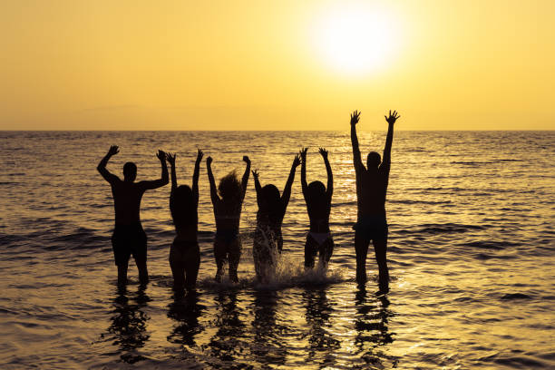 happy friends jumping inside water on the beach at sunset. young people having fun on summer vacation. travel, party and friendship concept. image - group of people women beach community imagens e fotografias de stock