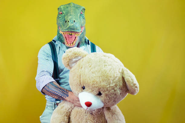 Senior man wearing t-rex dinosaur mask with teddy bear. Crazy man having fun celebrating carnival and valentin day. Absurd and surreal funny concept. Yellow background - Image