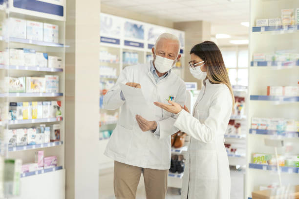 istock Education in pharmacy and corona virus. A mature pharmacist explains the documentation in the pharmacy to a young pharmacist in professional training. They wear uniforms and face masks 1318289326