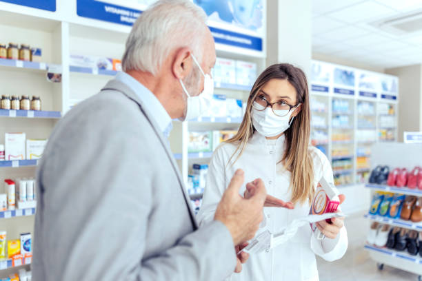 Help from a pharmacist. A female pharmacist with glasses holds medicine boxes and instructions for use. She looks at the man who is explaining something else. They both wear protective masks