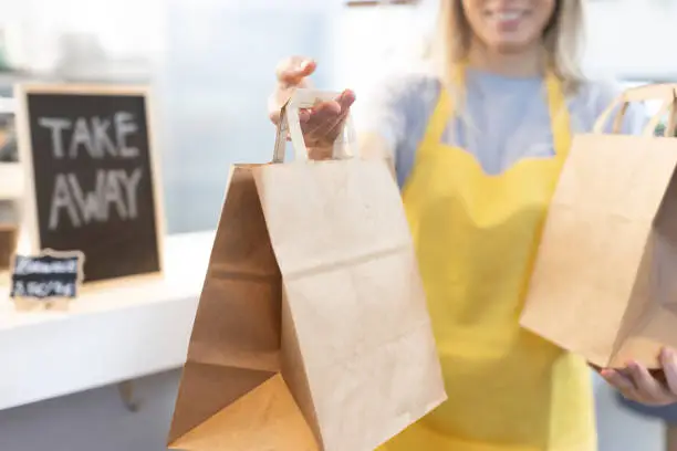 Photo of Young smiling blonde waitress in a yellow apron making a take away food delivery in recycled paper bags in a bar. Selective focus. Front view. Take Away concept.