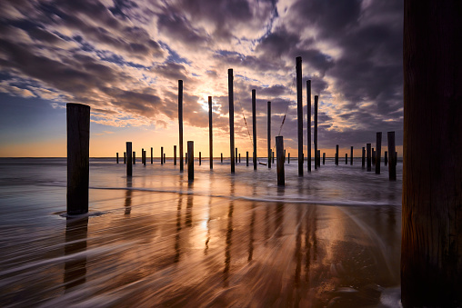 Poles on the beach in Petten during sunset. The sky is filled with dark clouds but the sun is shining brightly through. The poles create shadows on the water that is moving quickly.