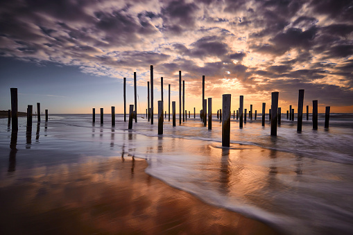 Poles on the beach in Petten during sunset. The sky is filled with dark clouds but the sun is shining brightly through. The poles create shadows on the water that is moving quickly.