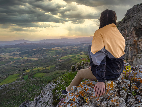 Woman in raincoat sitting on the edge of a cliff overlooking a valley on a rainy wet day. Conceptual outdoor tone image.