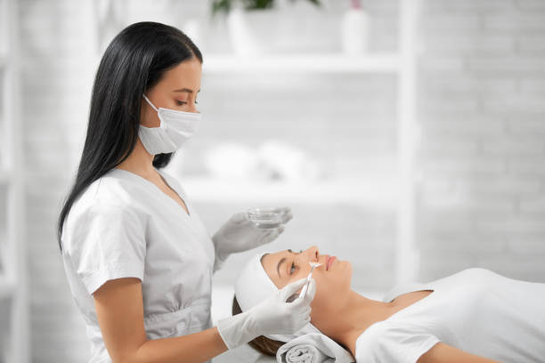 Young woman doing special procedure for improvements skin. Side view of young beautician in protective mask applying cream for young brunette patient woman. Concept of process peeling for improvements face skin. aesthetician photos stock pictures, royalty-free photos & images