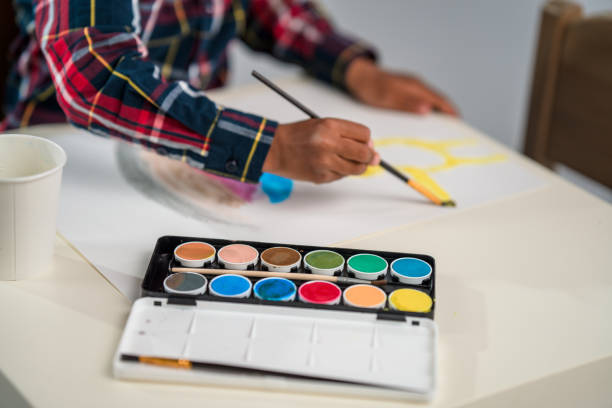 Mixed race child sitting behind a small table while painting a picture using a paint brush and colours.