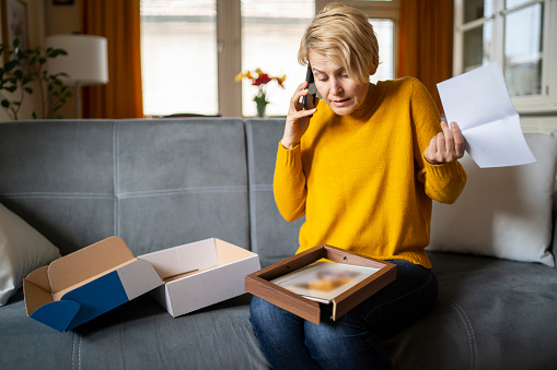 Angry confused woman sitting on the couch at home, looking at broken online store order is displeased by post shipping mistake and uses her phone to ring and complain