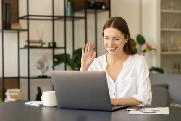 Photo of Smiling young Caucasian woman sitting at her office desk wave greet talk on video call online . Zoom conference concep
