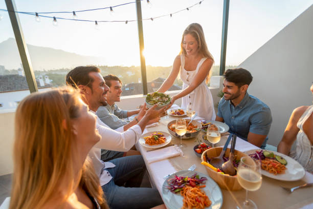 Friends and family passing salad on a table at sunset. Friends and family passing salad on a table at sunset. There is spaghetti Bolognese on some of the plates on the table. There are glasses of wine and other food on the table; everyone is having fun laughing and smiling passing giving photos stock pictures, royalty-free photos & images