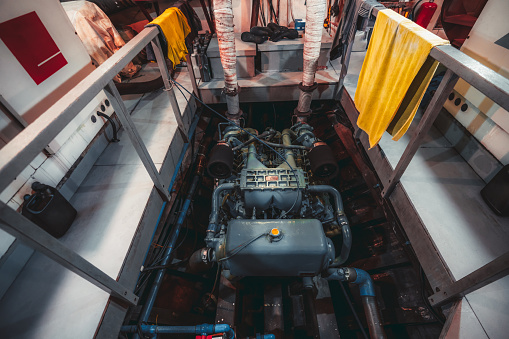 A wide-angle view of a huge powerful gasoline diesel engine of a yacht, or a boat, or a small ship, painted in blue and placed in a dark recess of an engine room of a vessel interior