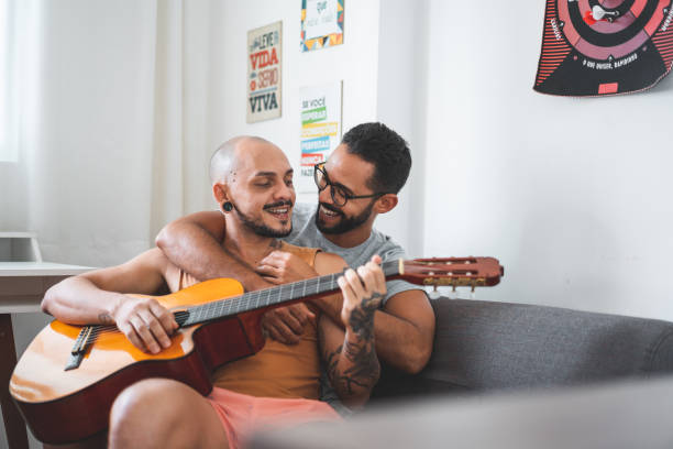 casal gay tocando guitarra e cantando no sofá - love embracing couple valentines day - fotografias e filmes do acervo