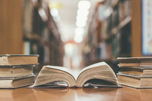 istock Book in library with old open textbook, stack piles of literature text archive on reading desk, and aisle of bookshelves in school study class room background for academic education learning concept 1318372912