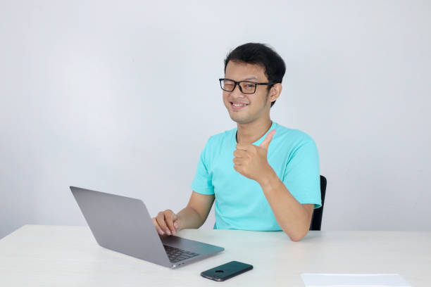 istock Young handsome asian man showing thumb up while working with laptop. Indonesian man wearing blue shirt. 1318386601