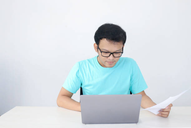 istock Young Asian Man is serious and focus when working on a laptop and document on the table. Indonesian man wearing blue shirt. 1318387435
