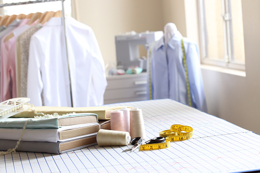 A pair of scissors and a tape measure, spools of thread, and several bolts of fabric rest on top of a pattern cutting board. A sewing machine and various tops hang from a rolling clothes rack out of focus in the background.