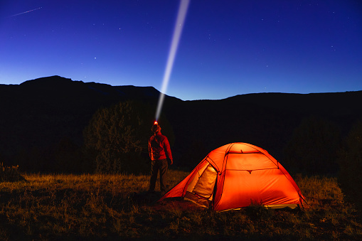 Hiker Camper Backpacker at Dusk with Glowing Orange Tent - Scenic mountain views during blue hour twilight of rugged backcountry setting. Man looking up at stars with headlamp projecting light beam into space up in sky.