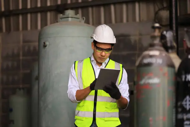 Asian industrial worker using digital tablet to check the coolant system in the factory while standing at liquid nitrogen and argon in compressed container tank for biochemical industry