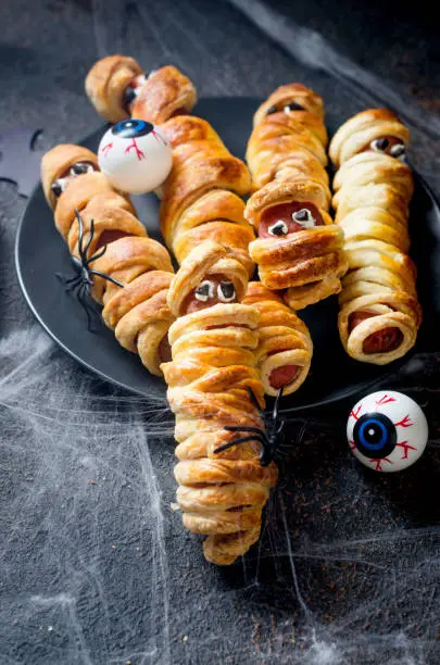 Spooky sausage mummies in dough in black plate with graves on dark table. Halloween funny food idea for kids party