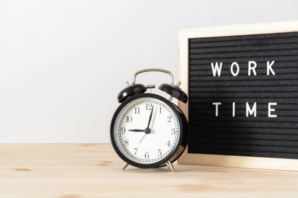 istock Classic black alarm clock on wooden table and black board with words 'work time' with white wall background and copy space. 1318397870