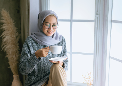 Confident Asian hijab woman enjoying a cup of coffee in the morning in front of windows and smiling