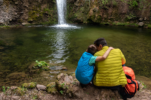 Dad and son child boy are happy together and embracing enjoy contemplation of waterfall lake among nature in mountains. Local travel, family vacations. Back view. Father's day concept