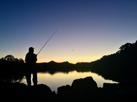 Horizontal landscape photo of a male fisherman, in silhouette, fishing in the tranquil waters of the Brunswick River, Brunswick Heads, near Byron Bay, NSW at dusk.