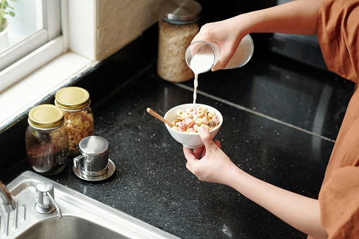 Woman pouring fresh milk in bowl of muesli and cornflakes for breakfast
