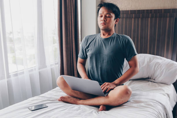 istock Work from home. Young Asian man sits on a bed. Home workplace. Break, relaxation at the time of work. 1318405982