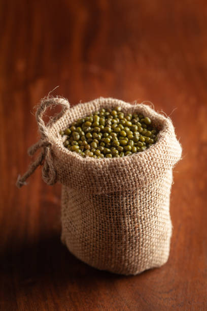 nahaufnahme von bio-grün gram (vigna radiata) oder ganz grün moong dal in einem stehenden jutebeutel über holzbraunem hintergrund. - mung bean stock-fotos und bilder