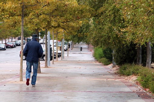 Older man with rain hat walking down the sidewalk seen from behind.