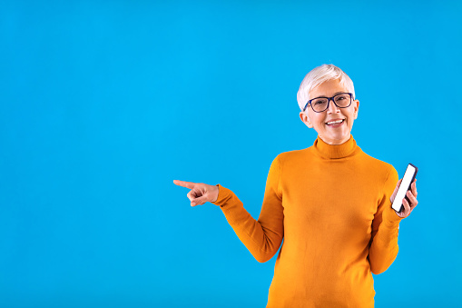 Senior woman, using mobile phone  while pointing on side against blue background