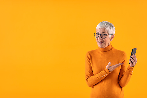 Studio portrait of an attractive mature woman using her phone against a yellow background, looking at camera.