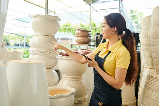 Young female greenhouse worker with smartphone in hands preparing big clay pots for selling in gardening center