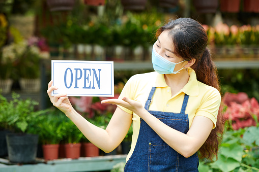 Young woman in medical mask opening plant nursery and showing sign to invite customers