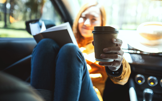 A young woman holding book and giving coffee cup in the car
