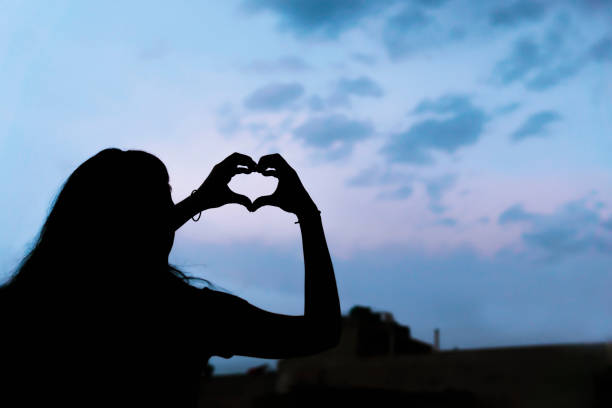 Low angle silhouette image of a young woman making heart shapes symbol with her hands against blue cloudy sky.