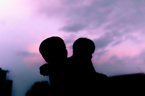Low angle silhouette image of two happy little brother enjoying and having fun together against blue and pink cloudy sky.