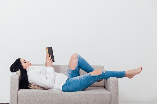 Relaxed young woman reading a book seated on sofa isolated on white background